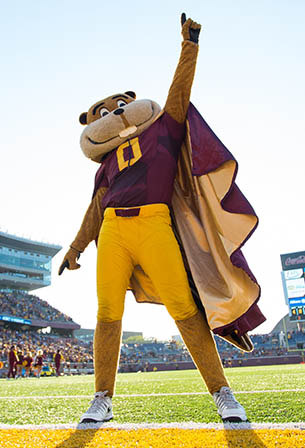 Goldy Gopher with cape standing on football field and raising arm air