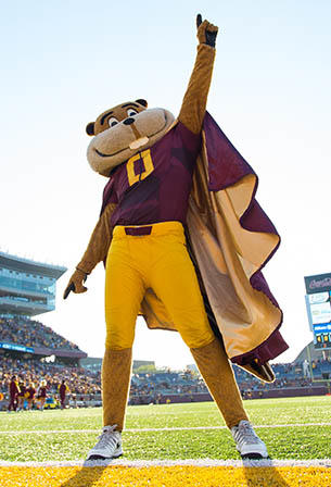 Goldy Gopher wearing a cape, pointing to the sky, and standing on a football field.